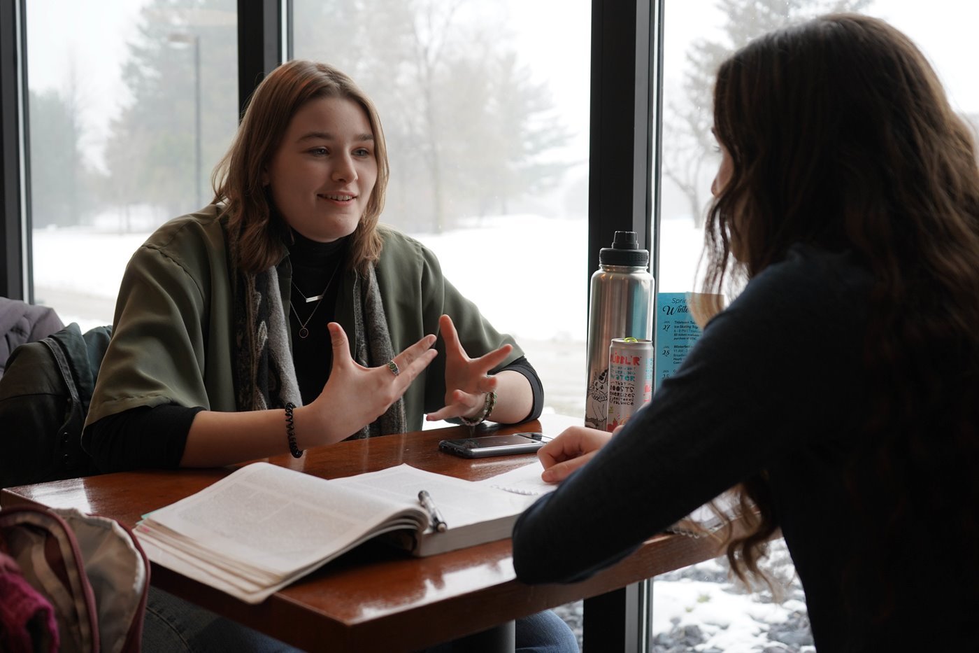 Two students sit at a table in front of a window with textbooks and water bottles between them