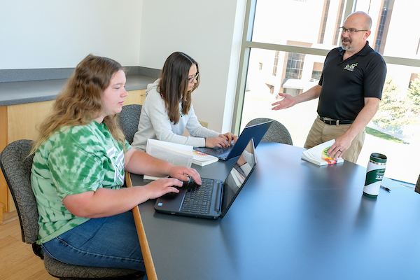 Two students sit at a table typing on their laptops. A professor stands in front of them with a book in his hand while explaining the course material.