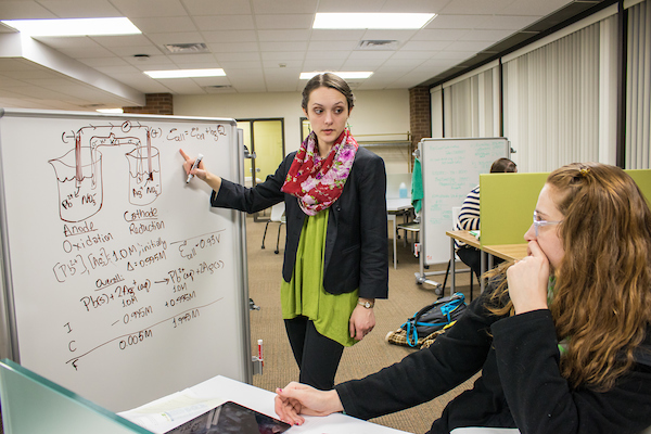 A tutor draws chemical diagrams and equations on a whiteboard while a student sits at a desk immediately in front of the board.