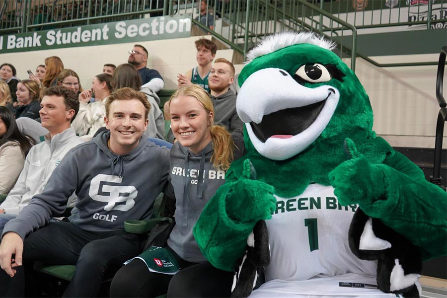 A group of people sitting in bleachers with a mascot  Description automatically generated