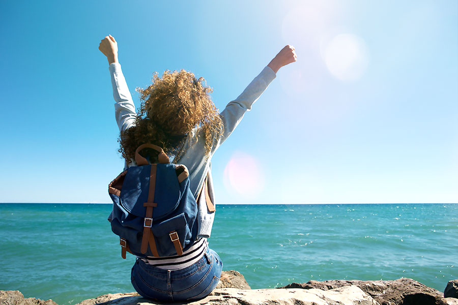 Young woman earing a backpack, arms raised jubiliantly facing the water