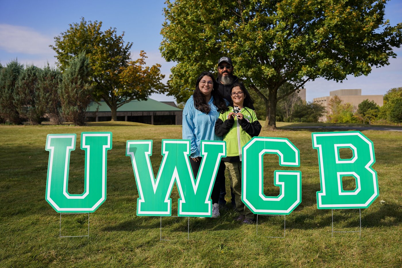 Three people post for a photo behind a "UWGB" yard sign