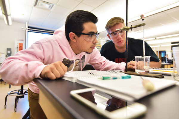 Two male students use ring stand, glass stirring rod and beaker